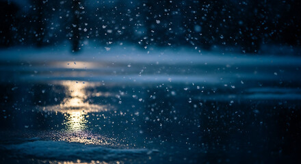 Fototapeta premium Gentle snowflakes fall softly onto a dark, reflective surface with a warm light shimmering in the distance, creating a serene winter atmosphere.
