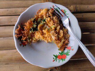 A close-up photo of a crispy Indonesian-style omelette served on a traditional floral-patterned enamel plate with a fork, placed on a bamboo table. The golden brown texture.