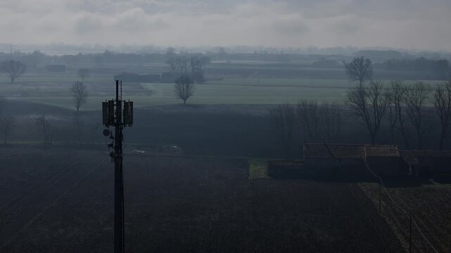 A rural cell tower stands amid fog-shrouded Italian farmlands, drone orbiting to capture its structure and the surrounding open fields, trees, and agricultural landscape on a misty morning.