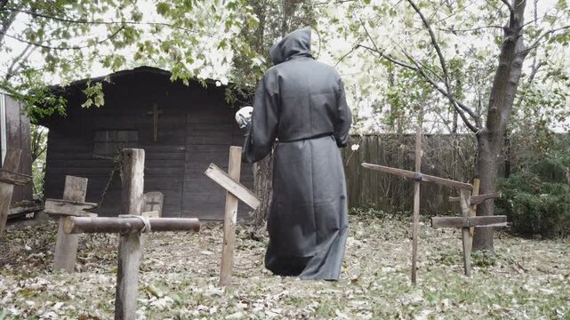 Grim reaper death in an old cemetery graveyard holding a skull and scythe with wood crosses during a rainy afternoon. Vintage classic horror or Halloween. Slider shot walking away in slow motion.