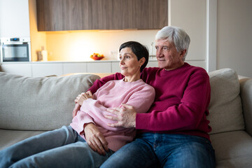 Relaxed senior spouses cuddle while enjoying watching TV or looking out window together. Happy elderly couple, relaxed retirement, affectionate seniors embracing on couch, elderly companionship