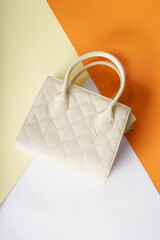 A cream-colored women's bag is placed against an orange, white, and cream background