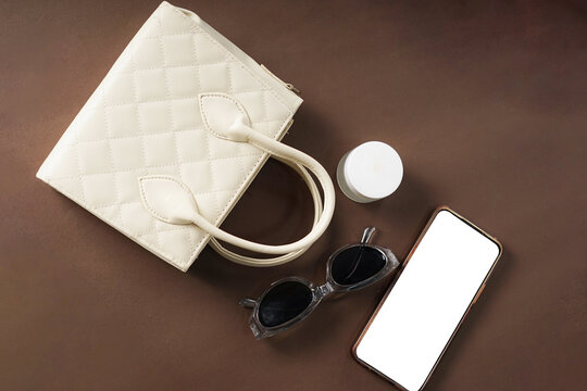 A cream-colored women's bag, sunglasses, a phone, and a moisturizer are placed against a brown background