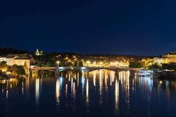 Fototapeta premium A peaceful cityscape at night shows a river reflecting bright city lights and a bridge. The scene highlights the beauty of illuminated buildings and calm waters, ideal for urban themes.