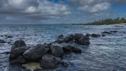 A pile of black volcanic boulders in the ocean. Ripples on the surface of the water. Green vegetation on the shore. Blue sky, clouds. Mauritius.