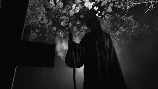 Classic grim reaper death in a cloak and holding a scythe with smoke or mysterious fog and backlighting in a cemetery graveyard. Edited in slow motion black and white.
