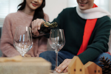 Couple wearing Santa hats celebrating Christmas at home, smiling while pouring red wine, surrounded by gifts and festive decorations.