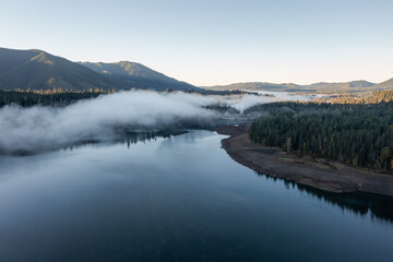 Aerial drone picture of sunrise at Wynoochee Lake dam in Olympic National Forest, Washington State, USA, showing calm water, low clouds, and forested mountain landscape in peaceful autumn morning	
