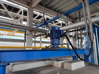 Top-down view of a blue industrial mixer motor and gearbox mounted on a processing tank in a chemical or manufacturing plant, surrounded by complex pipes and steel structures.