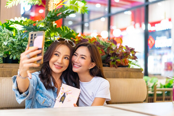 Happy Asian family mother and daughter using mobile phone taking selfie together. Adult child giving gift box to senior mother celebrating mother's day, birthday, Christmas or New Year holiday.