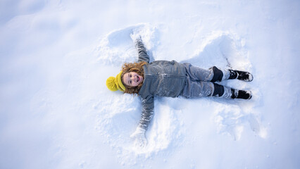 Fun winter day for a happy child. Snow angel made by a cheerful kid in winter. Child in cozy clothes lying in snow. Cute child making snow angels. Winter fun. Playful winter moment.