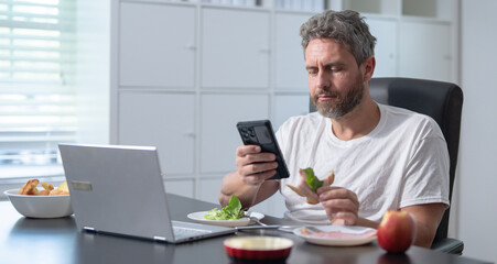 A casual office worker eats a breakfast while managing online tasks. Freelancer eating and working at a home office. Employee drinks starting his day with a healthy meal. Morning home office concept.