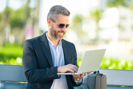 Confident business man sitting on a bench with a laptop and phone in park. Rich business man. Mature businessman enjoying work break. Businessman taking a break on a park bench. Summer business.