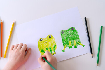 Child creates colorful frog drawings using crayons on a white sheet of paper in a bright, welcoming room
