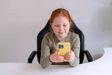Young girl sitting at a desk, smiling while using a yellow smartphone in a bright room during daytime