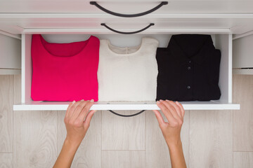 Young adult woman hands sorting folded different color classic shirts in white opened drawer box at...