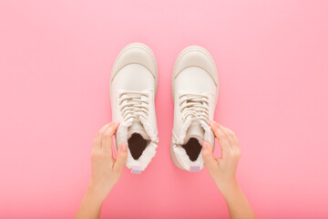 Little child girl hands holding and showing new white warm winter boots with shoelaces on light pink table background. Pastel color. Closeup. Point of view shot. Top down view.