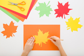 Child hands outlining contour with pencil for cutting maple leaf. Light gray table background. Creating colorful leaves from application paper. Making autumn decoration. Flat lay. Top down view.