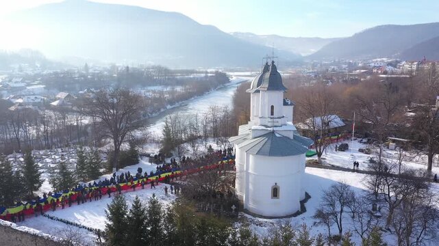 Winter aerial view of a crowd holding a large Romanian flag at Raducanu Monastery in Targu Ocna. The snowy town, river, and mountains are visible in the background.