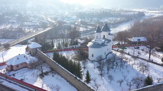 An ascending aerial shot slowly reveals Raducanu Monastery in Targu Ocna, Romania. A long line of people holds a massive Romanian flag on the snow-covered grounds during a winter national event.