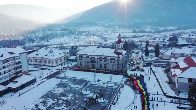 High-angle aerial shot of the snow-covered town square in Targu Ocna, Romania. A very long Romanian national flag is held by a line of people next to the historic town hall on a bright winter day.