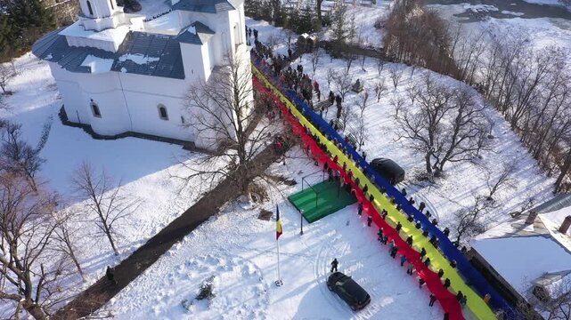 An ascending aerial drone shot of Raducanu Monastery in Targu Ocna, Romania, during winter. A long line of people holds a massive Romanian flag over the snowy grounds in a display of national pride.