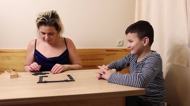 A woman and a boy are playing dominoes while sitting at a table.