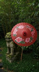 Ornate red paper parasol rests beside moss covered stone statue in lush green garden setting
