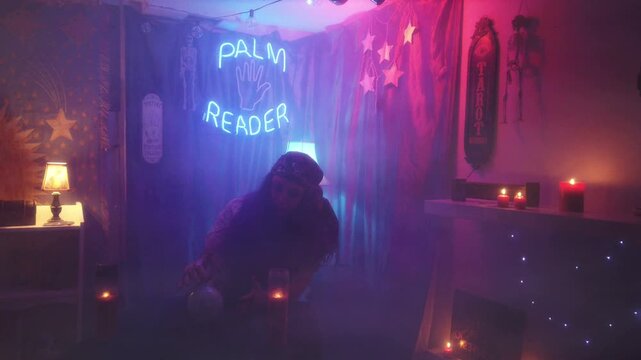 The interior of a psychic palm reader, fortune teller room, with a gypsy woman waving her hands over an electric crystal ball. With neon lights, candles and various spiritual materials. Handheld shot.