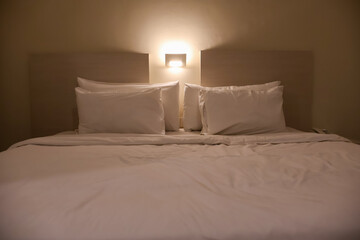 A comfortable, tidy hotel bed with crisp white linens and multiple pillows, featuring a light wooden headboard and a warm, low-intensity night light above.