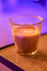 Close-up of a warm milk coffee beverage in a small glass cup with foam, illuminated by vibrant neon purple light in the background.