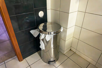 Metal trash bin in the corner of a public restroom, paper towels sticking out, white and black ceramic wall tiles and light tiled floor