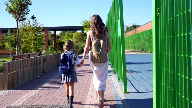 Mother and daughter holding hands while walking to school on a sunny day. The girl is wearing a uniform and carrying a backpack, representing the first day of school and family bonding