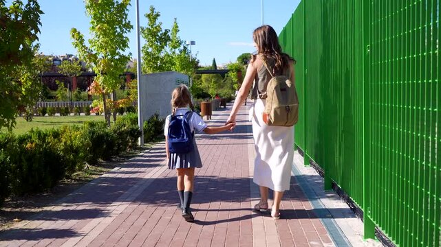 Young mother walking her little daughter wearing a uniform to school on a sunny day. A parent taking her child to elementary school, showing care, love, and support for her education