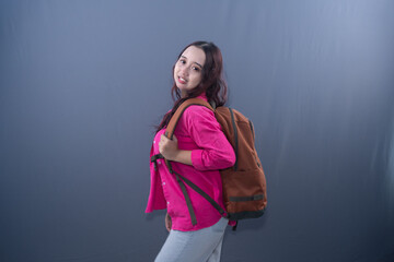 A smiling woman in a pink shirt and jeans wears a brown backpack, looking at the camera.