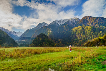 Traditional Tibetan woman standing in the wide green valley of Jiuzhaigou, China, surrounded by snow-capped mountains, autumn forest, and soft morning light under a blue sky.