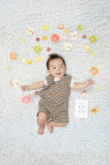 Naklejka premium A baby is laying on a white rug with a bunch of fruit around him. The fruit includes apples, oranges, and kiwis. The baby is wearing a brown and white outfit. The scene is playful and lighthearted