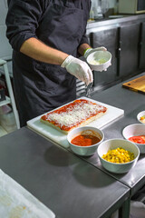 Anonymous male chef in gloves sprinkling grated cheese over a tomato sauced pizza dough on stainless steel counter, surrounded by bowls of fresh ingredients in a professional kitchen