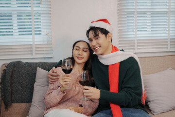 Happy couple wearing Santa hats enjoying wine together on the sofa beside a decorated Christmas tree, celebrating the festive holiday season at home.