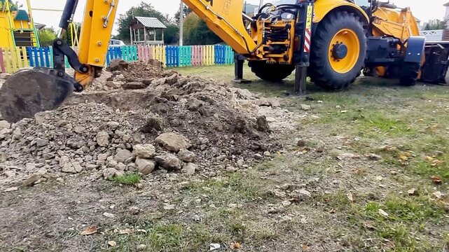 Yellow backhoe loader performs excavation work, digging soil with a bucket to create a trench. Demonstration of heavy construction machinery operation at a construction site during the day.