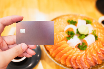 Hand holding a credit card in front of a large sashimi salmon platter at Japanese restaurant. Great for dining payment, gourmet lifestyle, seafood, fine dining, and modern food experiences.