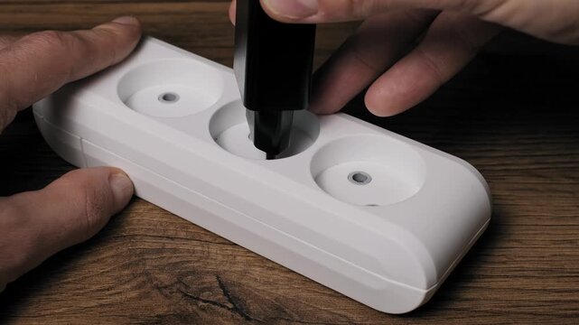 Angled shot of a male hand inserting a black European plug into a white triple outlet extension strip. The strip rests on a dark wood texture desk, connecting an electrical appliance to the grid.