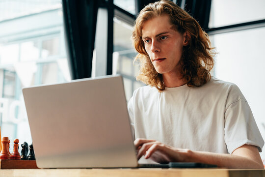 Young man working intently on laptop in modern cafe with chess pieces nearby - Powered by Adobe