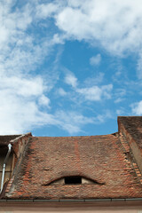 An old sloping roof with old tiles and a skylight, fragment of blue sky with clouds in the background.
