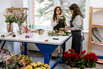 Two female florists creating beautiful rose bouquets together at bright modern flower shop workspace with professional floristry supplies and tools