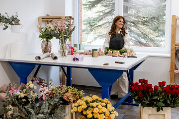 Happy female florist arranging fresh roses and flowers at workbench in bright modern flower shop studio with large windows and floral arrangements
