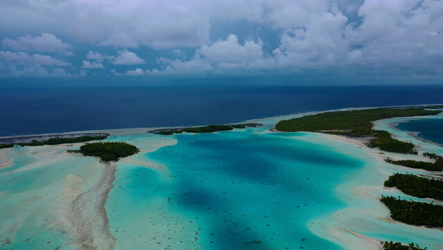 Aerial view of the bright turquoise lagoon contrasting with the dark blue ocean, fringed by lush green islets under a cloudy sky, Rangiroa, Tuamotu Islands, French Polynesia.