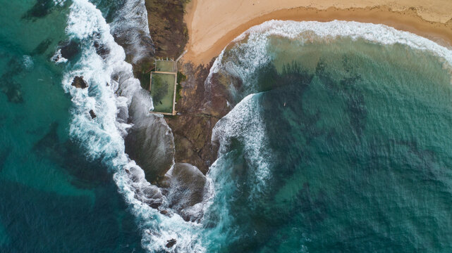 Aerial view of turquoise waves crashing against the shore and tidal pool, contrasting with the sandy beach's warm tones, creating a mesmerizing coastal scene, Sydney, New South Wales, Australia.
