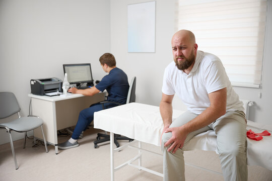 Male patient being examined in clinic for knee pain. Male suffering from knee pain osteoarthritis. Close up man holding his knee due to physical injury while sitting in room.