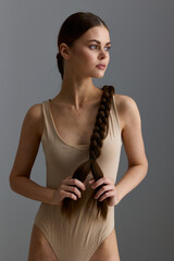young woman with long braided hair wearing beige bodysuit standing against gray background, natural light, minimalistic style, thoughtful expression